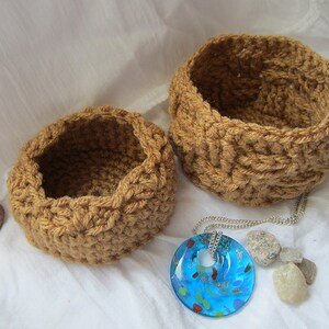 May include: Two brown crocheted baskets of different sizes, with a blue glass pendant necklace on a white surface.