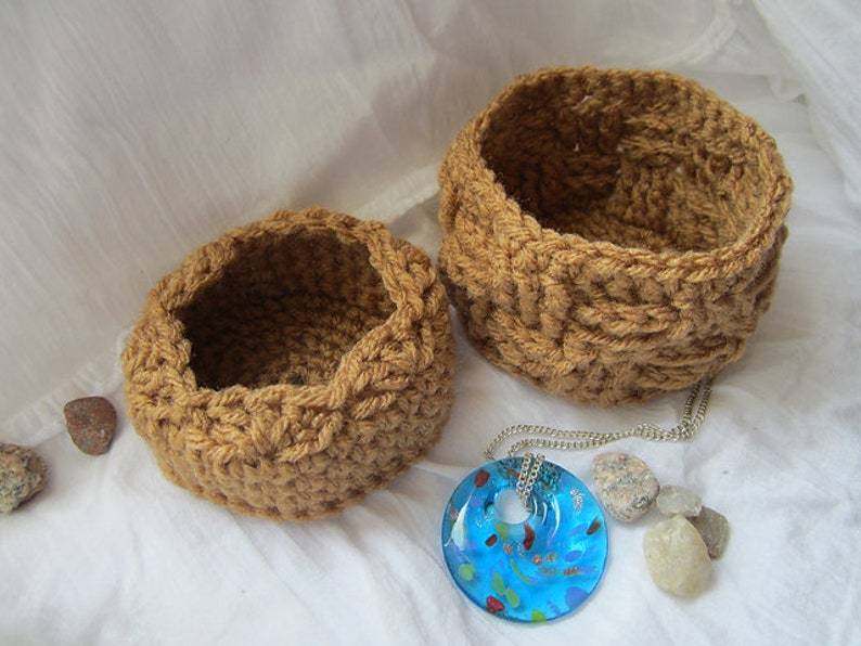 May include: Two brown crocheted baskets of different sizes, with a blue glass pendant necklace on a white surface.