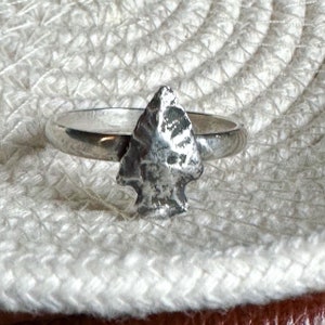 May include: A silver ring featuring a detailed arrowhead design. The ring has a polished band and a textured arrowhead centerpiece. The ring is displayed on a white, textured surface.