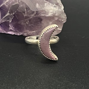 May include: A silver ring with a purple crescent moon shaped stone. The stone has a textured edge.