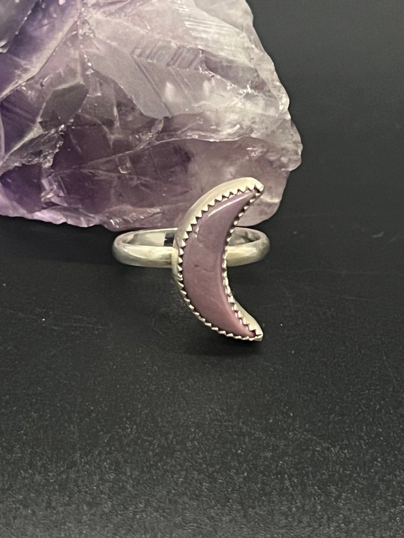 May include: A silver ring with a purple crescent moon shaped stone. The stone has a textured edge.