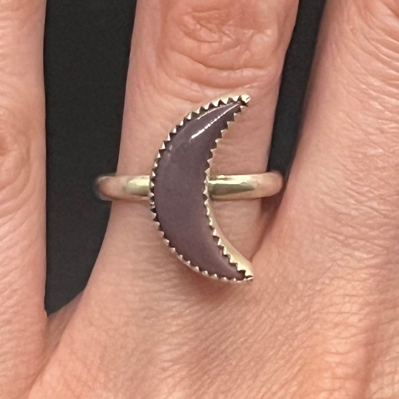 May include: A silver ring with a purple crescent moon shaped stone. The stone is set in a silver band with a scalloped edge.