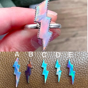 May include: A silver ring with a lightning bolt shaped opal gemstone. The opal is iridescent with pink, blue, and purple hues. The ring is shown on a hand wearing a silver band.