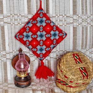 May include: A red and blue embroidered square cushion with a tassel hanging from the bottom. The cushion is hanging on a wall with a woven texture.