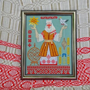 May include: Framed cross-stitch artwork depicting a figure in traditional dress, holding a sun and a dove. The artwork has a light blue background with red, orange, and white details. The frame is gold-coloured.