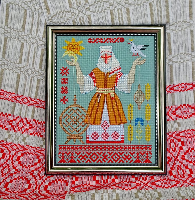 May include: Framed cross-stitch artwork depicting a figure in traditional dress, holding a sun and a dove. The artwork has a light blue background with red, orange, and white details. The frame is gold-coloured.