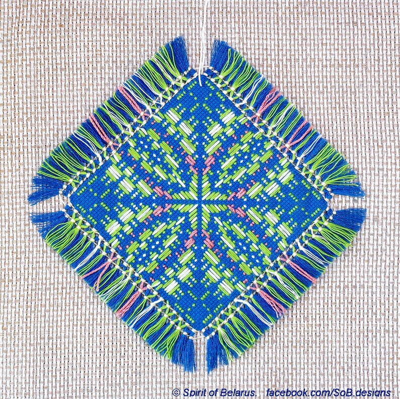 May include: A blue, green, white, and pink square-shaped embroidered ornament with fringe on all sides. The ornament features a geometric pattern with a central cross.
