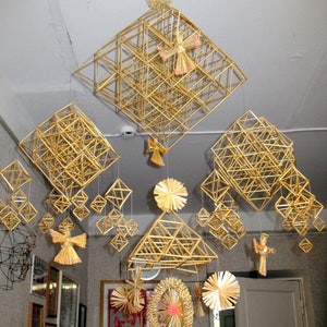 May include: A collection of straw ornaments hanging from the ceiling. The ornaments are made of woven straw and are in the shape of stars, angels, and geometric shapes. The ornaments are a light gold color and are hanging from white string.