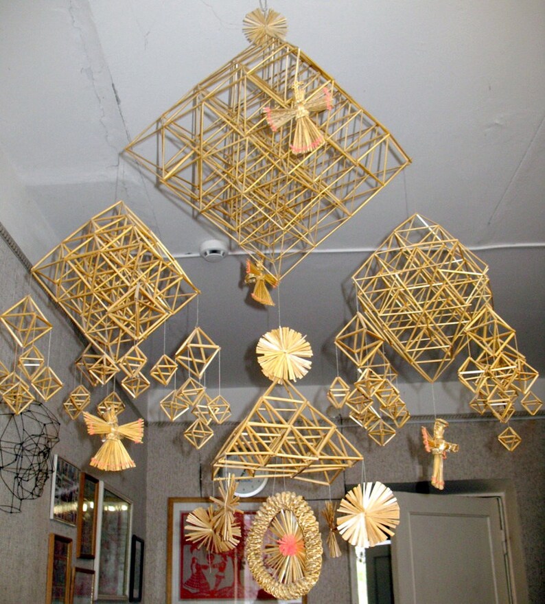May include: A collection of straw ornaments hanging from the ceiling. The ornaments are made of woven straw and are in the shape of stars, angels, and geometric shapes. The ornaments are a light gold color and are hanging from white string.
