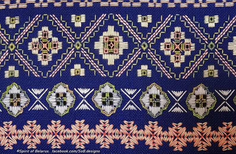 May include: Close-up of blue fabric with detailed embroidery. The design features geometric patterns in white, green, pink, and gold. The embroidery includes floral and diamond shapes, creating a textured surface.
