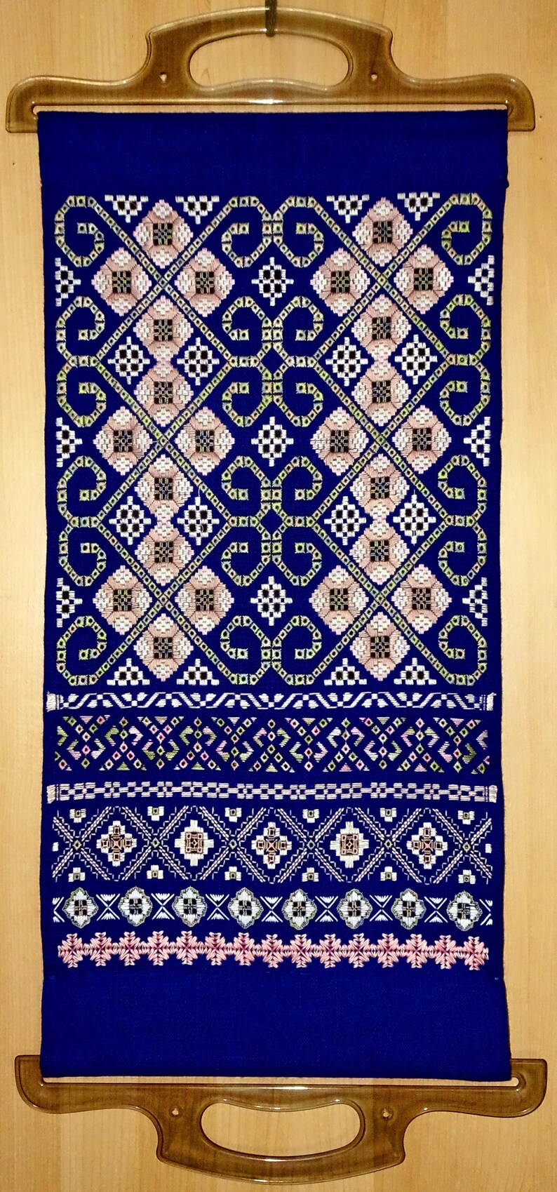 May include: A blue textile with detailed geometric patterns in white, pink, and brown. The design features repeating diamond and square shapes, with a decorative border. The textile is displayed with a wooden hanger.