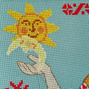 May include: Cross-stitch embroidery showing a smiling sun holding a flower, with a hand reaching up to meet it. The design is on a light blue background, with red and white accents. The embroidery is in progress, with visible needle and thread.