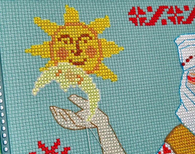 May include: Cross-stitch embroidery showing a smiling sun holding a flower, with a hand reaching up to meet it. The design is on a light blue background, with red and white accents. The embroidery is in progress, with visible needle and thread.