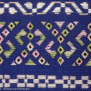 May include: Close-up of a blue textile with detailed embroidery. The design incorporates geometric patterns in white, pink, and green thread. The top and bottom borders feature repeating geometric shapes.
