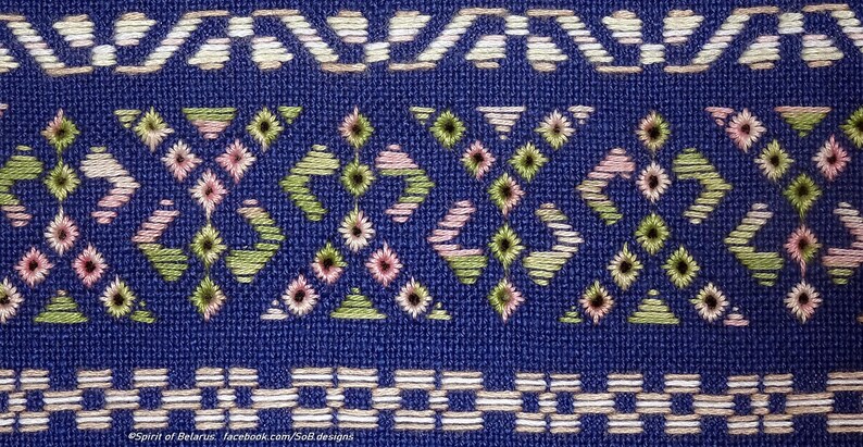 May include: Close-up of a blue textile with detailed embroidery. The design incorporates geometric patterns in white, pink, and green thread. The top and bottom borders feature repeating geometric shapes.
