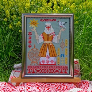 May include: Framed cross-stitch artwork depicting a woman in traditional Belarusian dress. The design incorporates a sun, bird, and folk symbols in red, yellow, and white on a light blue backdrop. The frame is silver.