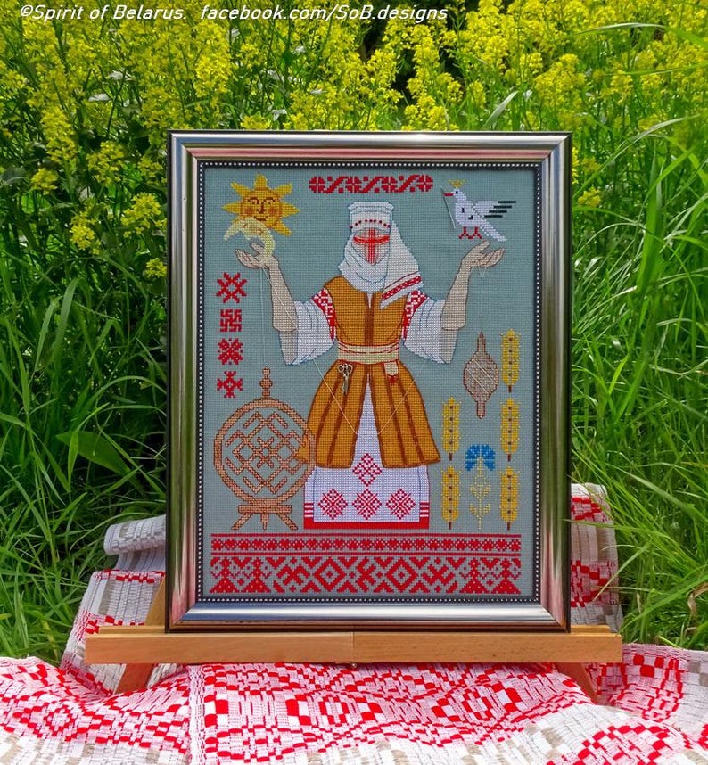 May include: Framed cross-stitch artwork depicting a woman in traditional Belarusian dress. The design incorporates a sun, bird, and folk symbols in red, yellow, and white on a light blue backdrop. The frame is silver.