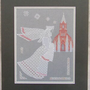 May include: White cross-stitch embroidery of an angel with wings, wearing a white dress and veil, flying towards a red church building with a steeple. The background is a gray fabric with white snowflakes.