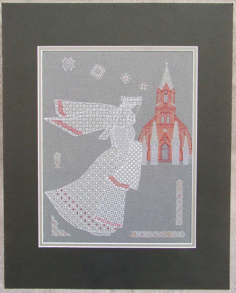 May include: White cross-stitch embroidery of an angel with wings, wearing a white dress and veil, flying towards a red church building with a steeple. The background is a gray fabric with white snowflakes.