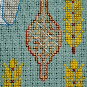 May include: Close-up of a cross-stitch design on a light blue fabric. The design features a central, elongated diamond shape in shades of orange and red, with a white thread overlay. Surrounding the central shape are yellow and red cross-stitched figures.