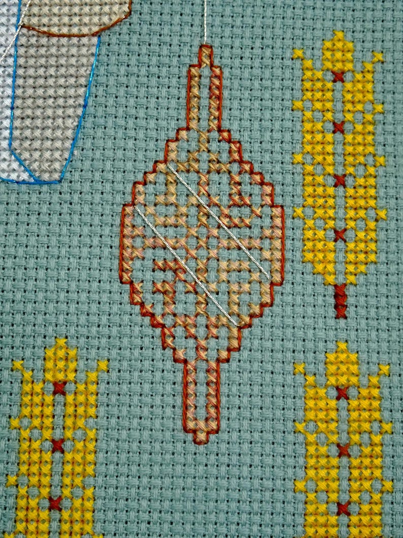 May include: Close-up of a cross-stitch design on a light blue fabric. The design features a central, elongated diamond shape in shades of orange and red, with a white thread overlay. Surrounding the central shape are yellow and red cross-stitched figures.
