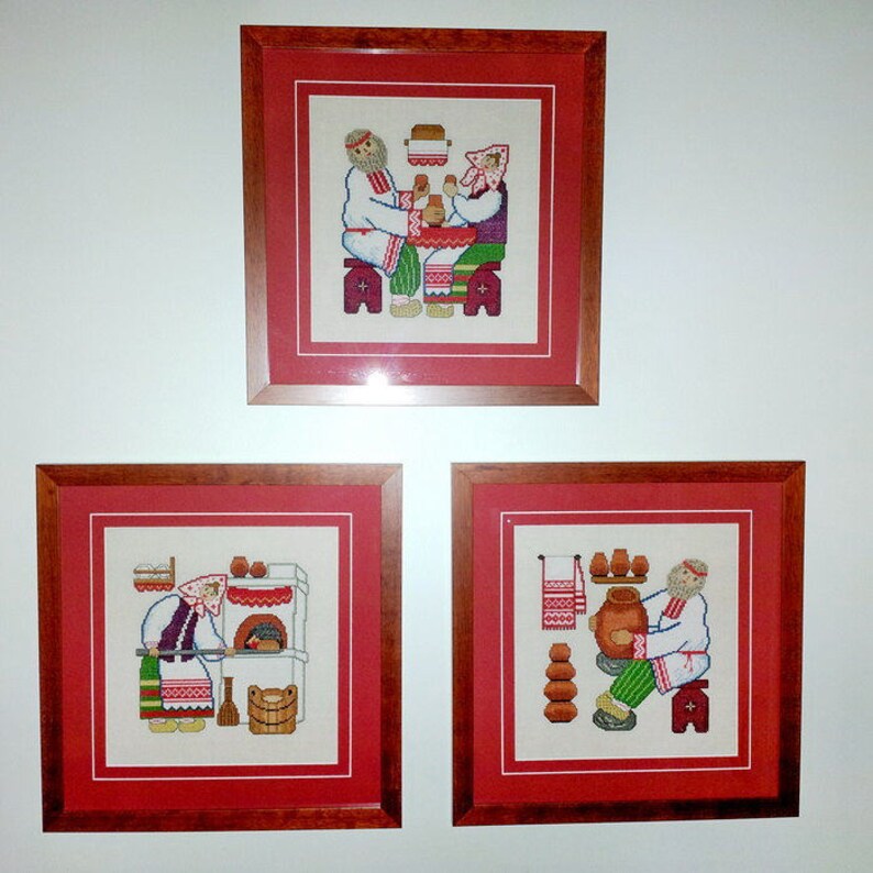 May include: Three framed cross-stitch embroidery pieces depicting scenes of traditional Russian life. The first piece shows a man and woman sitting at a table, the second shows a woman baking bread in an oven, and the third shows a man making pottery.