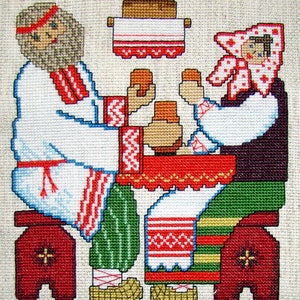 May include: Cross-stitch embroidery depicting a man and woman in traditional clothing sitting at a table with a loaf of bread. The man is wearing a white shirt with red and white embroidery, and the woman is wearing a white shirt with red and white embroidery and a red and white headscarf.