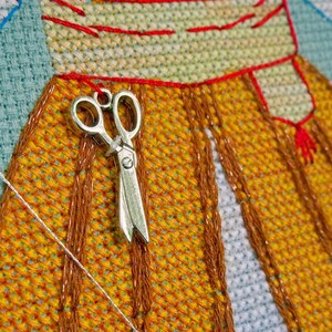May include: A silver pair of scissors hangs on a cross-stitch project. The project has a yellow and orange pattern with red and brown stitching. A white thread and blue fabric are also visible in this close-up shot.
