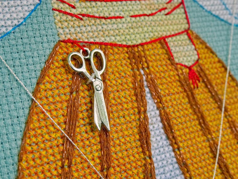 May include: A silver pair of scissors hangs on a cross-stitch project. The project has a yellow and orange pattern with red and brown stitching. A white thread and blue fabric are also visible in this close-up shot.