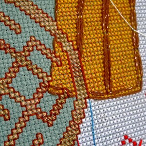May include: Close-up of cross-stitch embroidery showing geometric patterns in blue, gold, and red. The detailed stitching is visible, highlighting the texture of the fabric. The craft is in progress, with threads and unfinished areas.