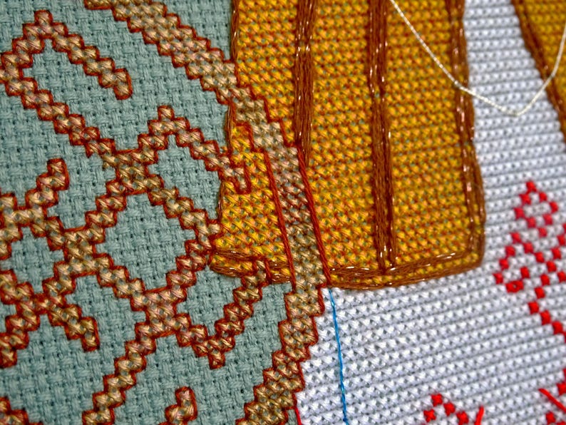 May include: Close-up of cross-stitch embroidery showing geometric patterns in blue, gold, and red. The detailed stitching is visible, highlighting the texture of the fabric. The craft is in progress, with threads and unfinished areas.