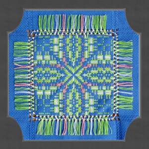 May include: A blue square with a white, green, and pink cross-stitch design. The design is surrounded by a fringe of white, green, and pink threads.