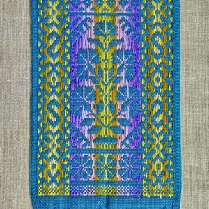 Op de afbeelding: Een blauwe geweven wandhanger met een geometrisch patroon in geel, paars en roze. De hanger heeft een franje van blauw, geel en paars garen.
