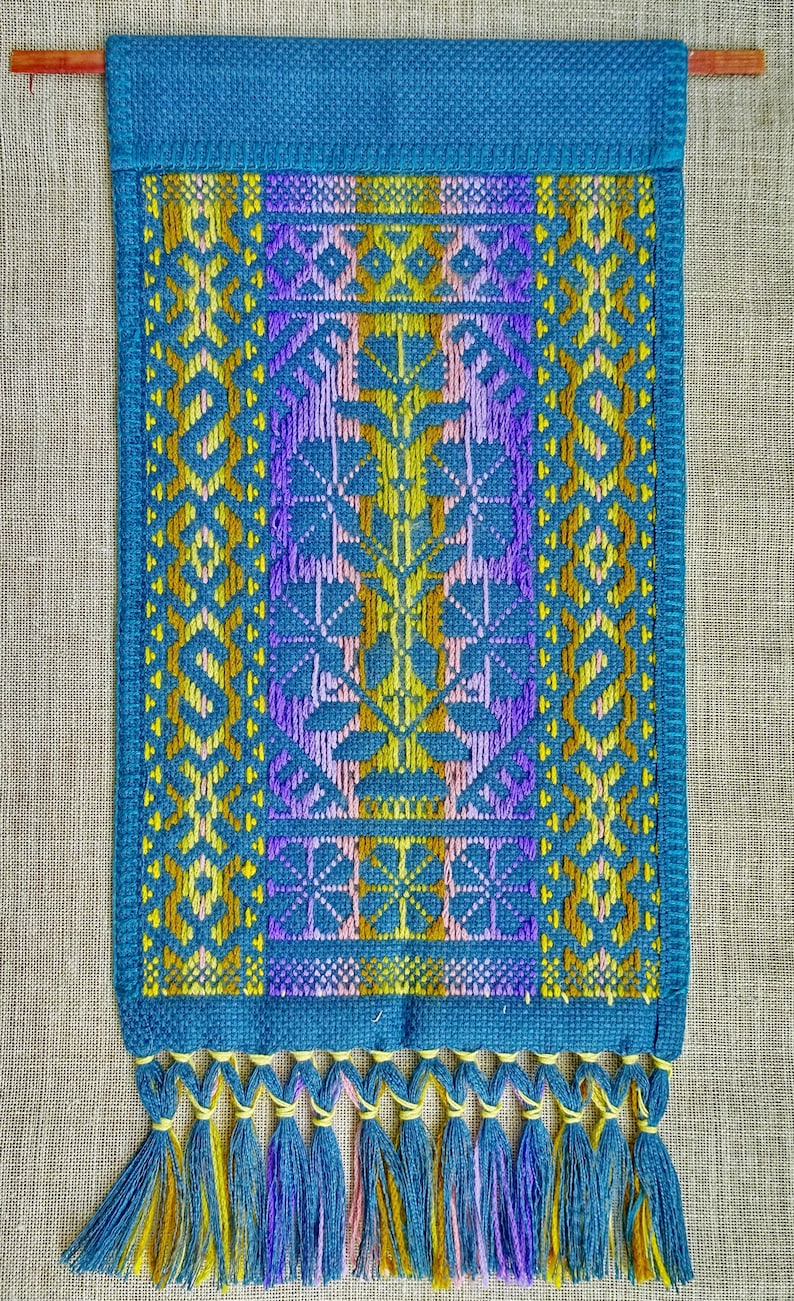 Op de afbeelding: Een blauwe geweven wandhanger met een geometrisch patroon in geel, paars en roze. De hanger heeft een franje van blauw, geel en paars garen.