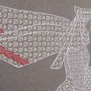 May include: White cross-stitch embroidery on a gray linen fabric. The design features a woman in a white dress and a red ribbon. The woman is standing in front of a building.