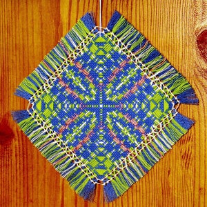 May include: A square, blue, green, and white crocheted doily with fringe hanging from a wooden surface. The doily has a geometric pattern in the center.