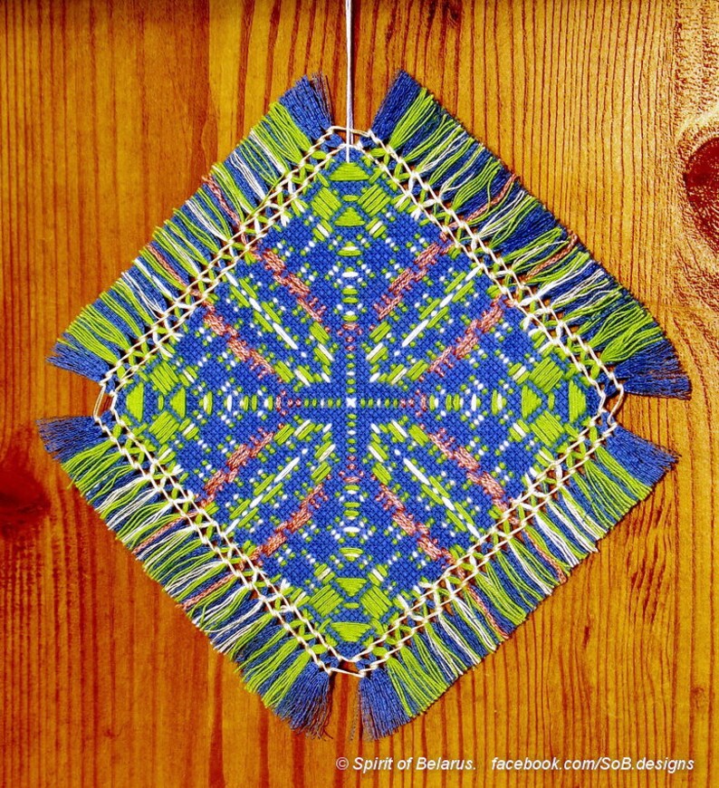 May include: A square, blue, green, and white crocheted doily with fringe hanging from a wooden surface. The doily has a geometric pattern in the center.