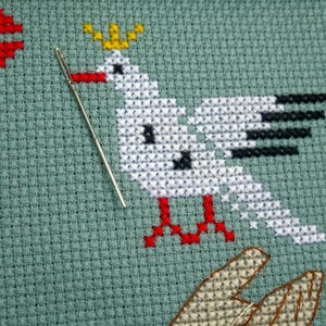 May include: A cross-stitch design on green fabric depicts a white bird with black wing details, red feet, and a yellow crown. A silver needle is positioned near the bird. Additional red and brown cross-stitch elements are visible.