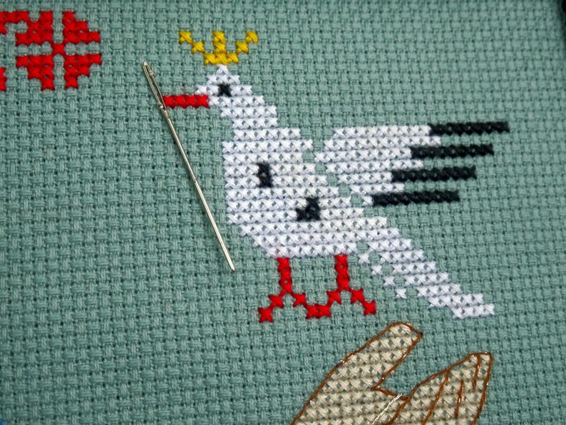 May include: A cross-stitch design on green fabric depicts a white bird with black wing details, red feet, and a yellow crown. A silver needle is positioned near the bird. Additional red and brown cross-stitch elements are visible.