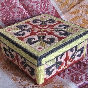 Könnte beinhalten: Eine kleine quadratische Schachtel mit einem bunten geometrischen Muster, das auf der Oberseite und den Seiten gestickt ist. Die Schachtel ist aus cremefarbenem Stoff mit schwarzen und roten Akzenten.
