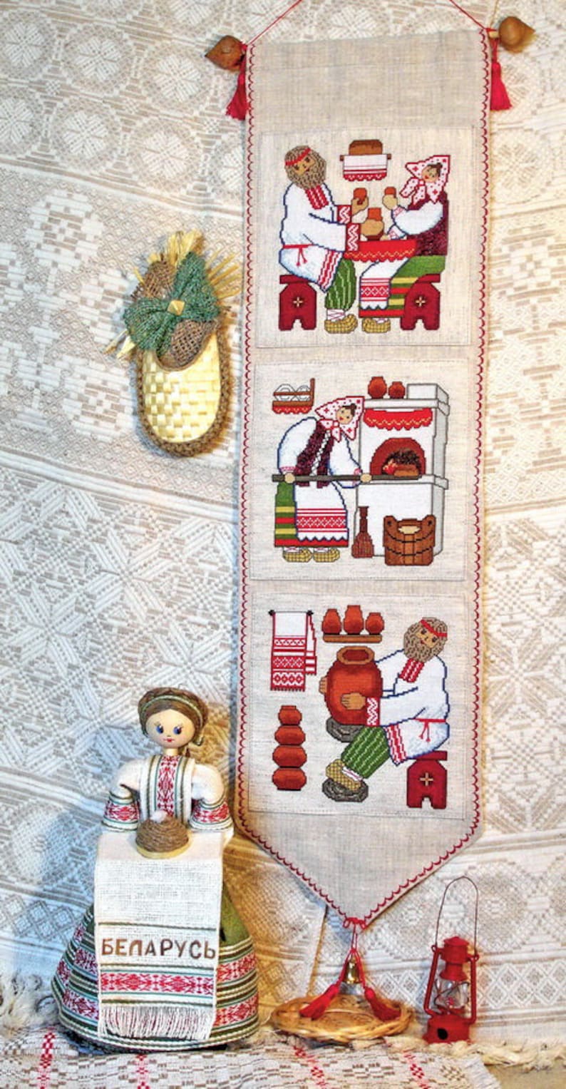 May include: A white linen tapestry with three embroidered scenes of people in traditional Belarusian clothing. The scenes depict a couple sitting at a table, a woman baking bread, and a man making pottery. The tapestry is hanging on a wall with a white and brown patterned background. The word "BELARUS" is embroidered on a doll in the foreground.