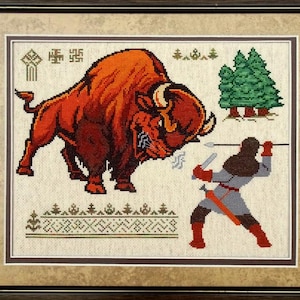 K&ouml;nnte beinhalten: Ein gerahmtes Kreuzstich-Stickbild, das einen gro&szlig;en, rotbraunen Bison zeigt, der einer Figur mit einem Speer gegen&uuml;bersteht. Die Szene enth&auml;lt gr&uuml;ne Kiefern, geometrische Muster und kleine Symbole auf hellem Stoff, eingefasst in einem dunklen Rahmen.