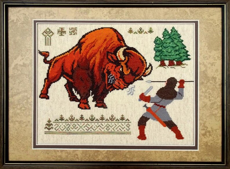 K&ouml;nnte beinhalten: Ein gerahmtes Kreuzstich-Stickbild, das einen gro&szlig;en, rotbraunen Bison zeigt, der einer Figur mit einem Speer gegen&uuml;bersteht. Die Szene enth&auml;lt gr&uuml;ne Kiefern, geometrische Muster und kleine Symbole auf hellem Stoff, eingefasst in einem dunklen Rahmen.