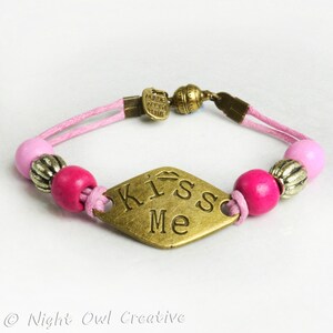 May include: A pink and gold bracelet with a diamond-shaped charm that says "Kiss Me".