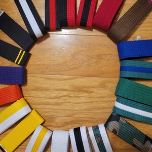 May include: A collection of martial arts belts arranged in a circle on a wooden surface. The belts are in various colors, including black, white, red, blue, green, brown, and yellow.