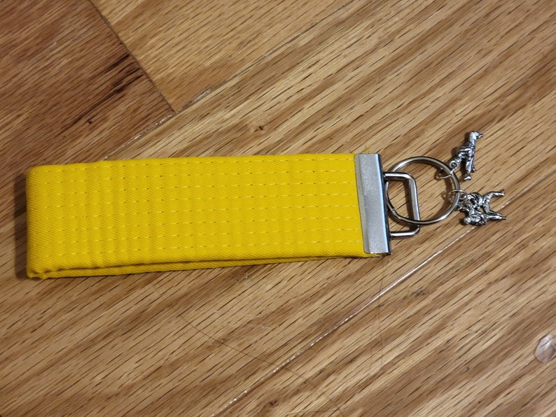 May include: A yellow karate belt keychain with a silver key ring and two silver charms. The charms are of a person doing a karate move and a person doing a martial arts pose.