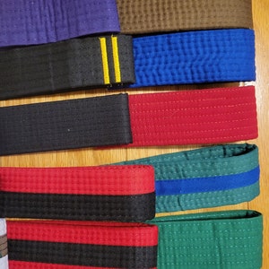 May include: A collection of martial arts belts in various colors, including red, black, blue, green, and orange. The belts have different colored stripes and are arranged in a row on a wooden surface.
