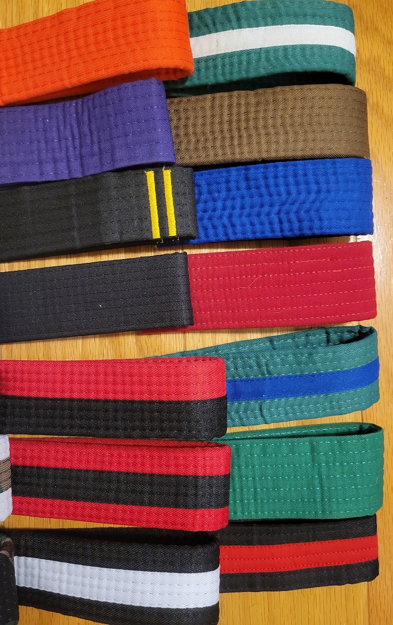 May include: A collection of martial arts belts in various colors, including red, black, blue, green, and orange. The belts have different colored stripes and are arranged in a row on a wooden surface.