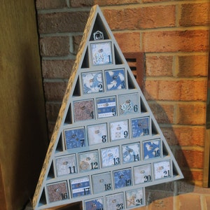 May include: A blue wooden Christmas tree advent calendar with 24 numbered drawers. Each drawer has a different design with a winter theme. The numbers 1 through 24 are visible on the drawers.