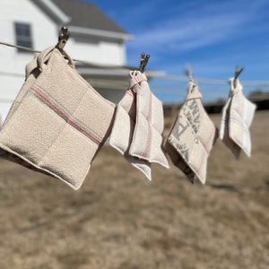 May include: Four beige pot holders with red stripes and vintage-style text hang on a clothesline. The pot holders are clipped to the line with metal clips. The background includes a white house and a clear blue sky.